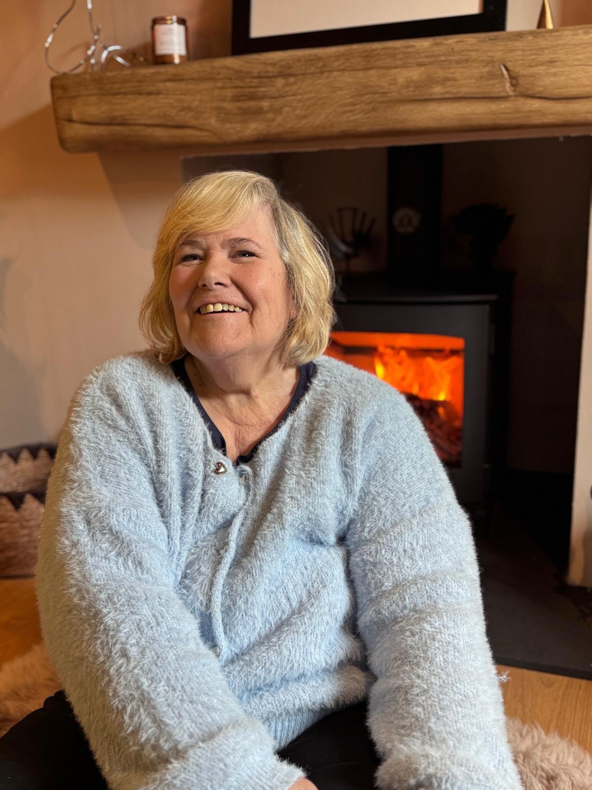 Jeanie Gother smiling warmly in her home beside a glowing fireplace